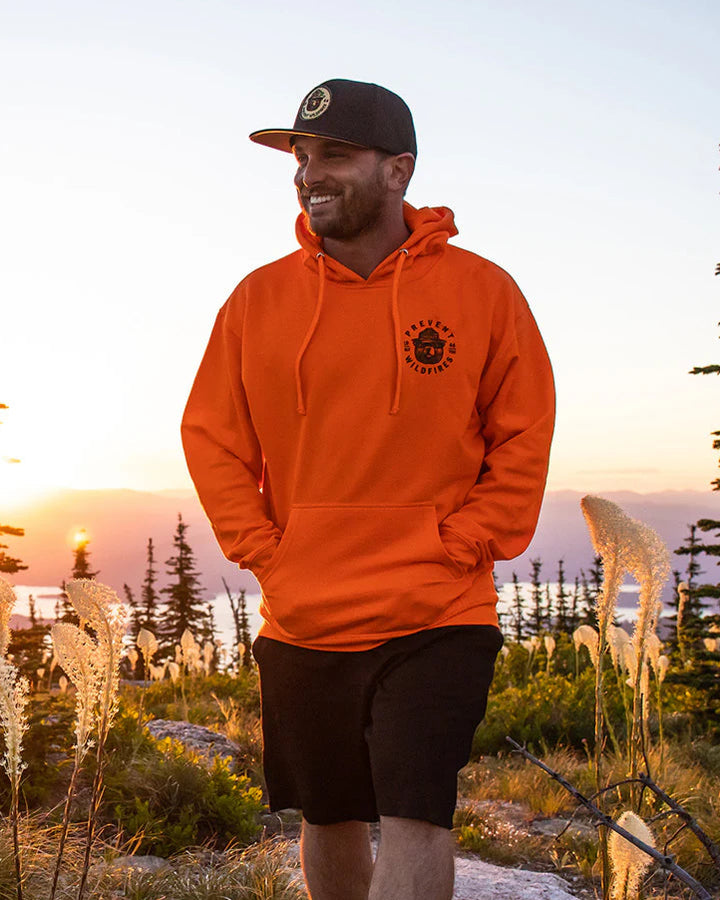 Man in an orange hoodie standing in a natural setting with trees and sunset.