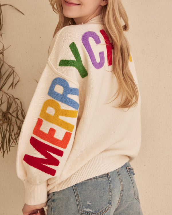 Person wearing a cream sweater with colorful letters spelling 'MERRY CHRISTMAS' on a beige background.