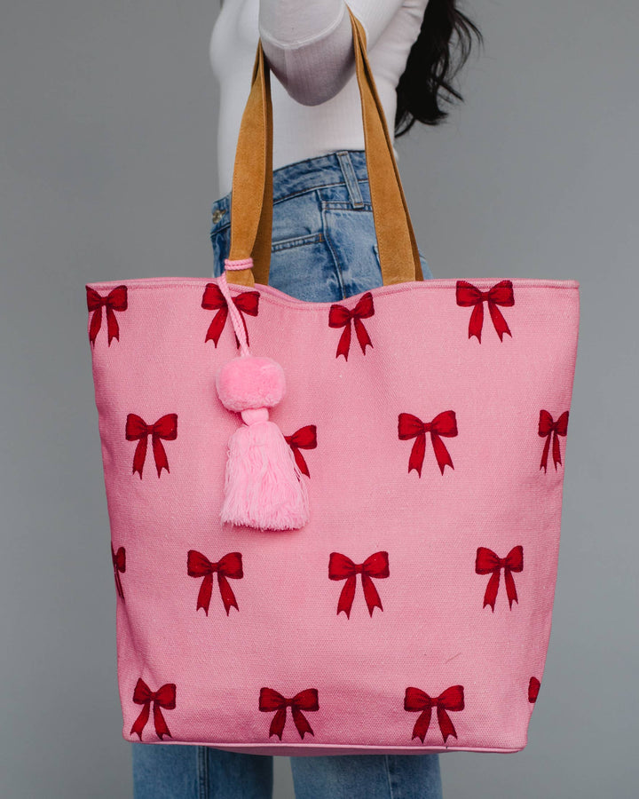 Pink tote bag with red bows and a pink pom-pom held by a person against a gray background