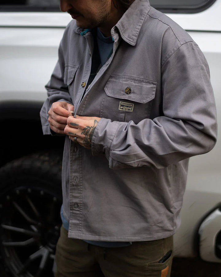 Man wearing a gray jacket with a logo, standing in front of a white vehicle.